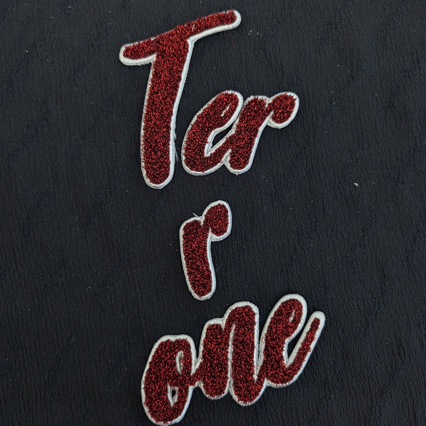 Red glitter "Terrone" patch with white outline on a dark fabric background.