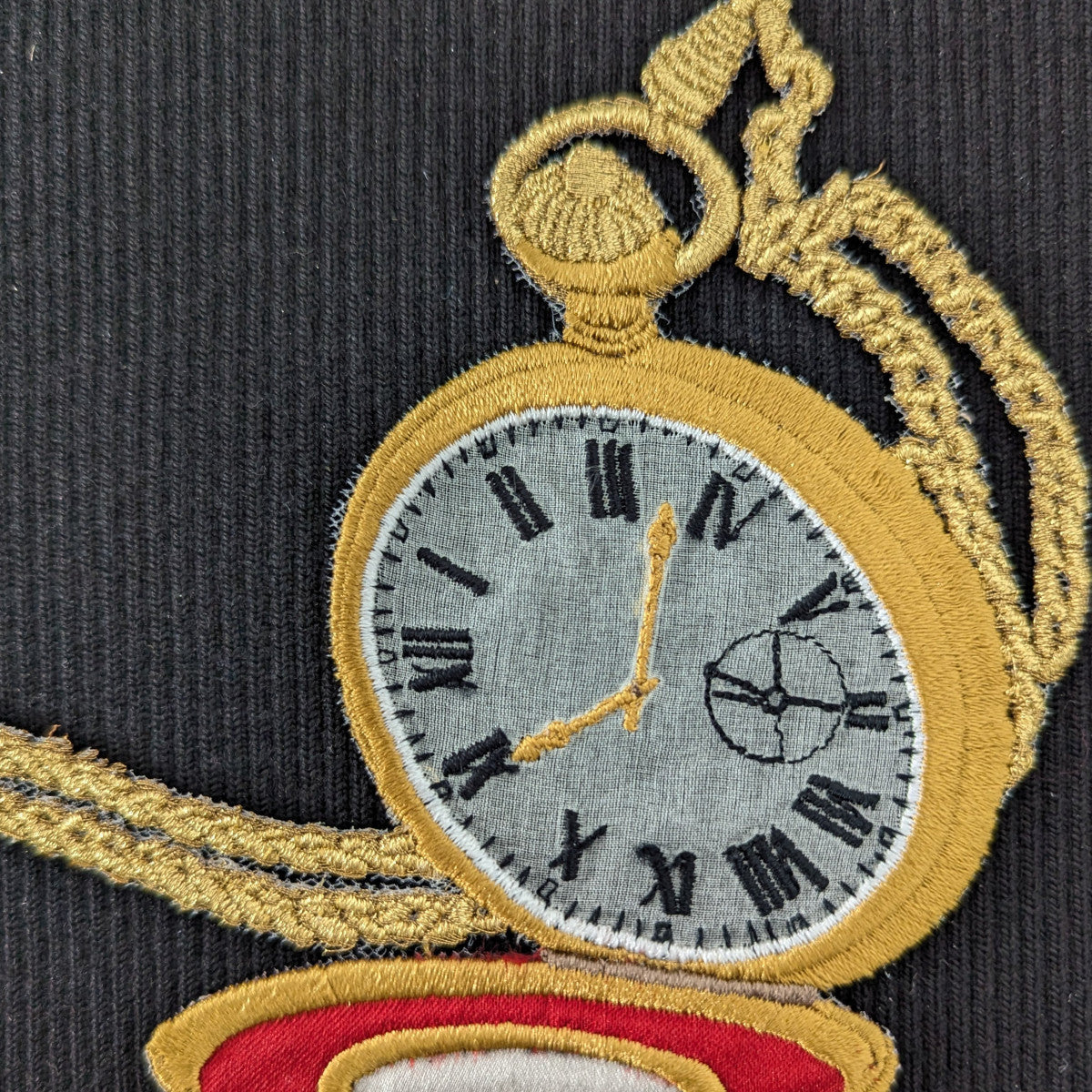 Gold embroidered pocket watch patch with gray face, black Roman numerals, and chain on black fabric.