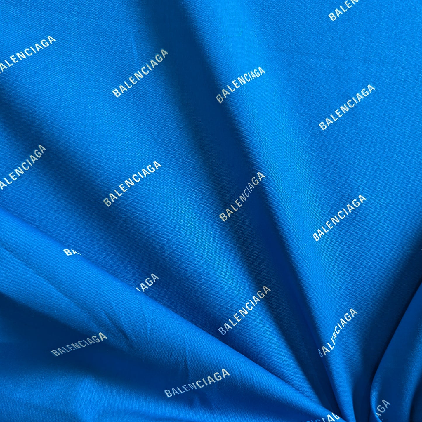 Bright blue cotton poplin fabric with repeating white "BALENCIAGA" logo print, draped.