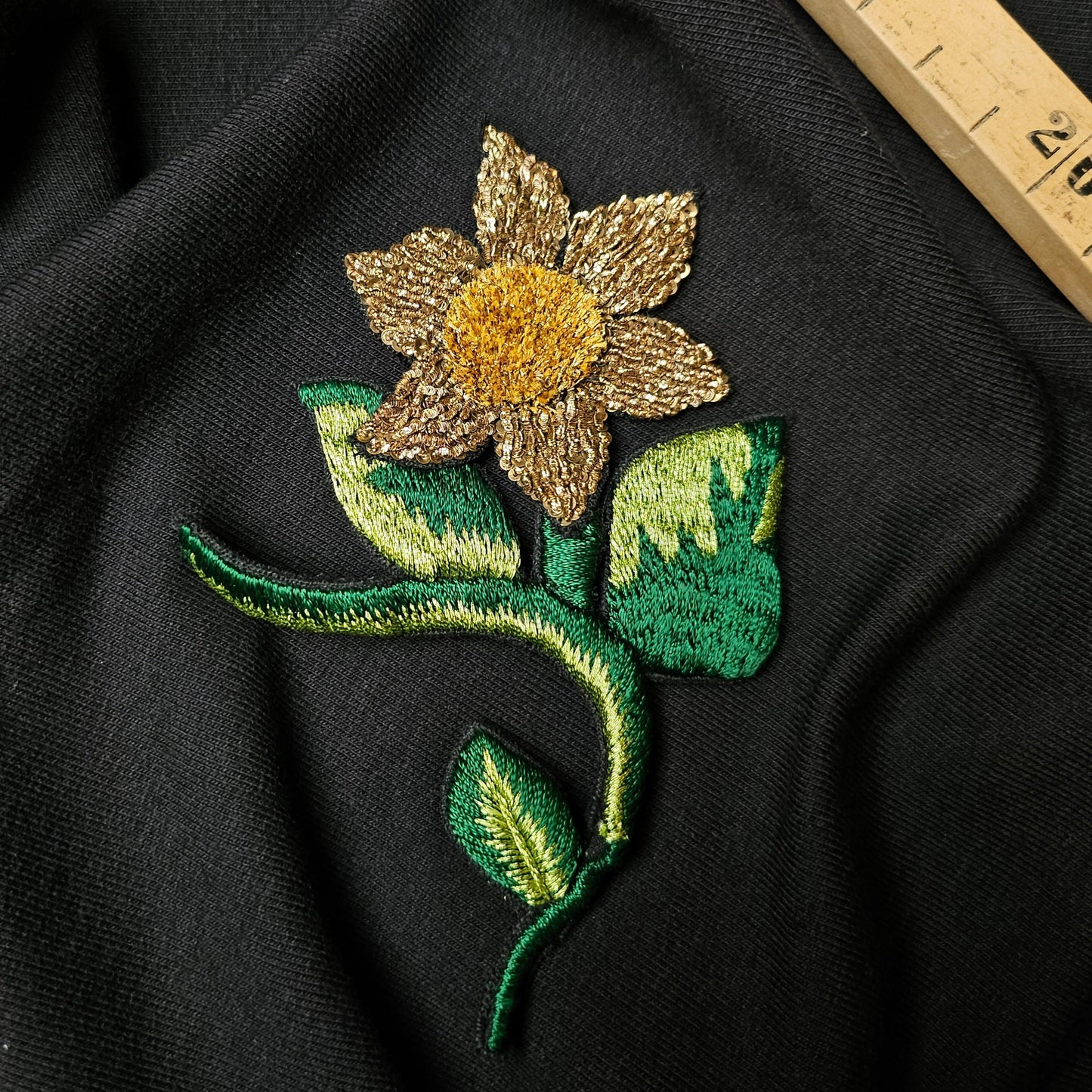 Embroidered D&G flower patch, 12cm, with gold petals and green leaves on black fabric.