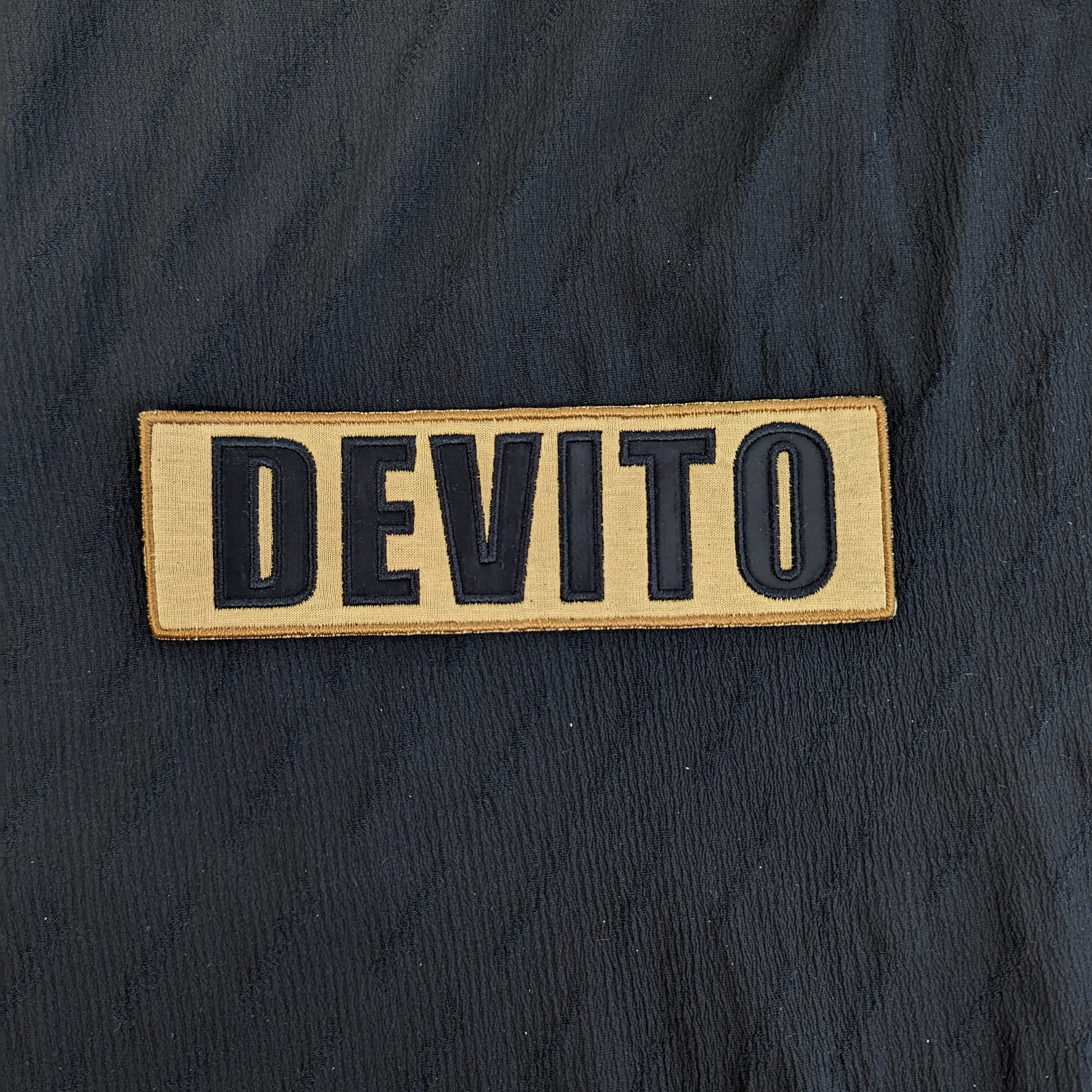 Embroidered "DEVITO" patch, 17x5.5cm, with black letters on a tan mixed fabric background.