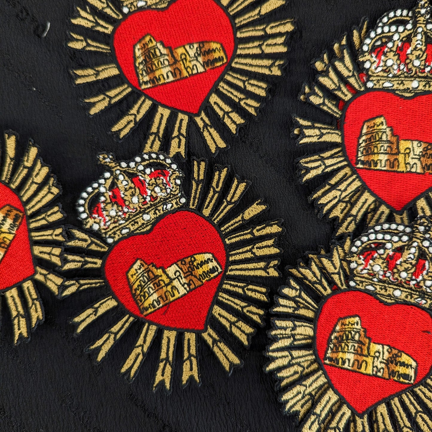 Red heart patches with gold rays, crowns, and Colosseum embroidery on black fabric.