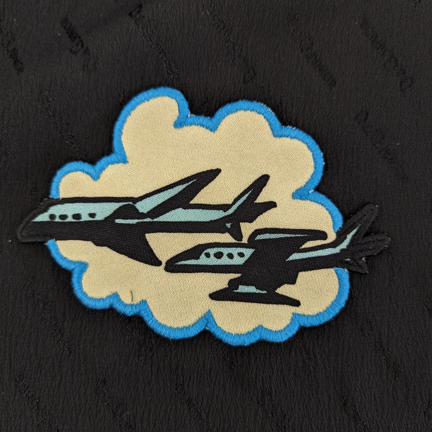 Black, tan, and blue DG Flugzeuge patch with two airplanes on a cloud, 13x9cm.