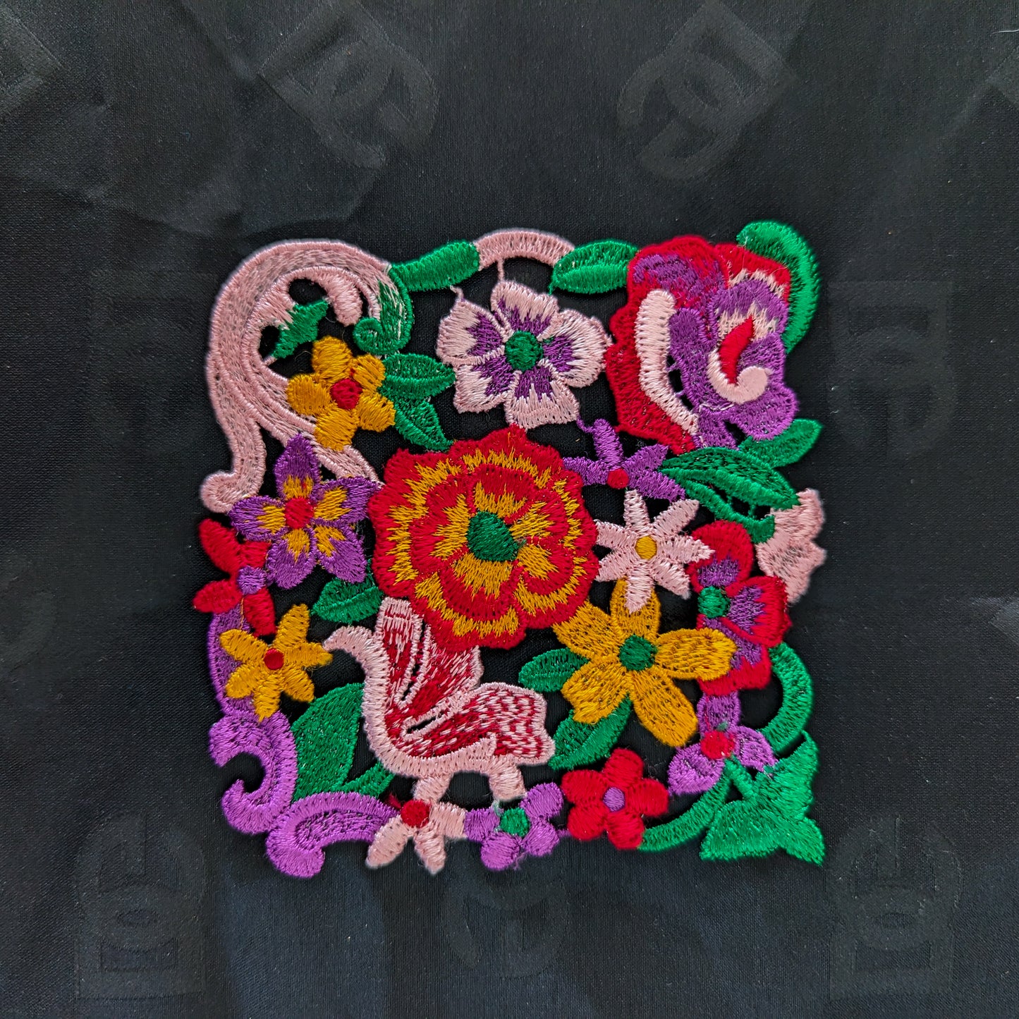 Colorful embroidered floral patch with a bird design, about 12 cm square, on a black background.