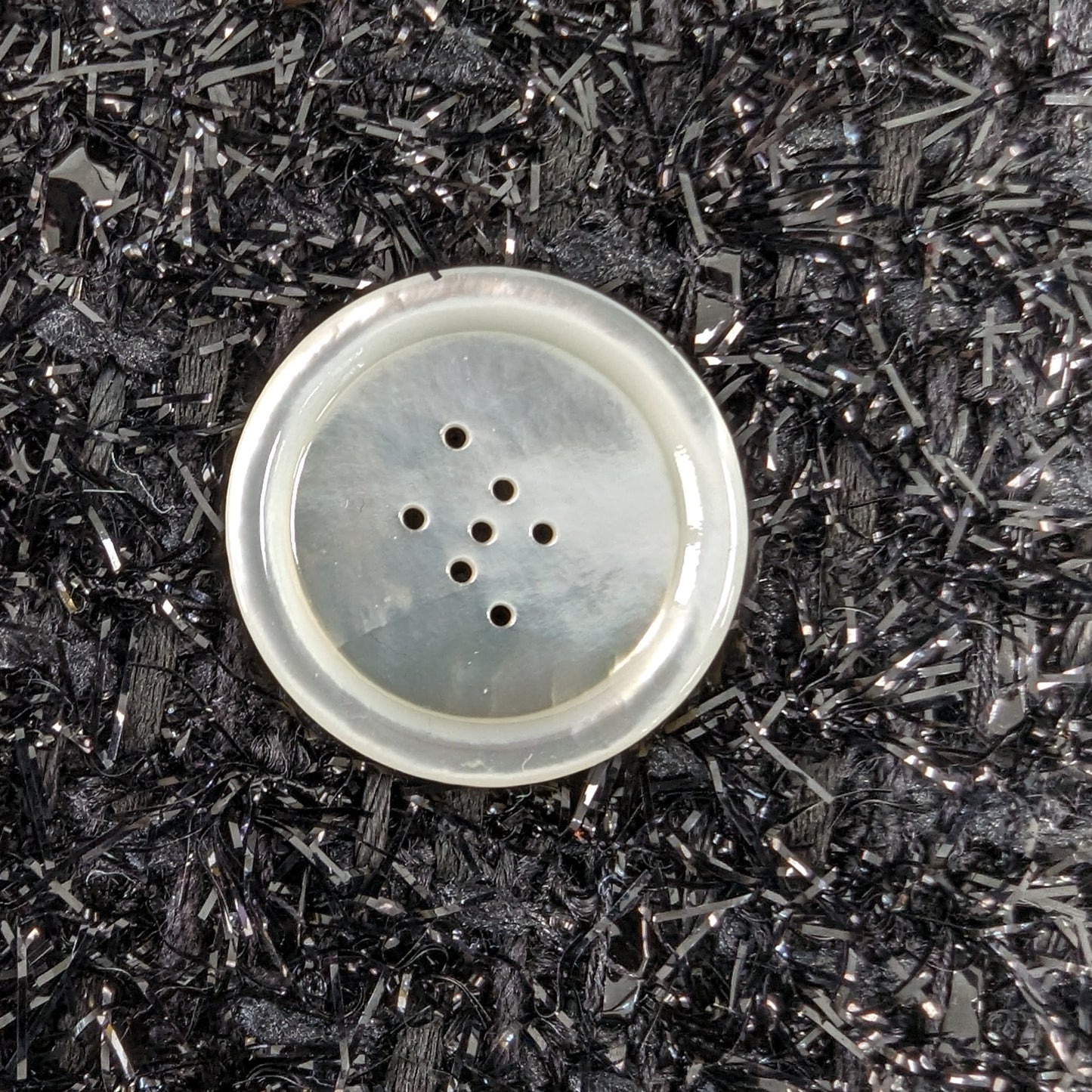 Iridescent mother-of-pearl button, 2.5cm, with 7 holes, on a black glitter background.
