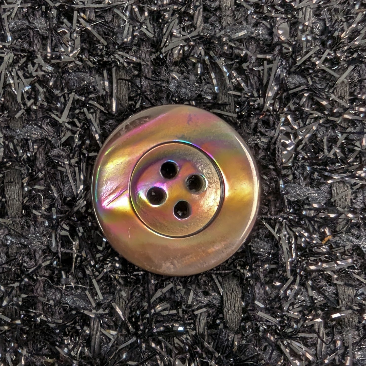 Iridescent mother-of-pearl button with four holes and a rim, on a dark, textured fabric.