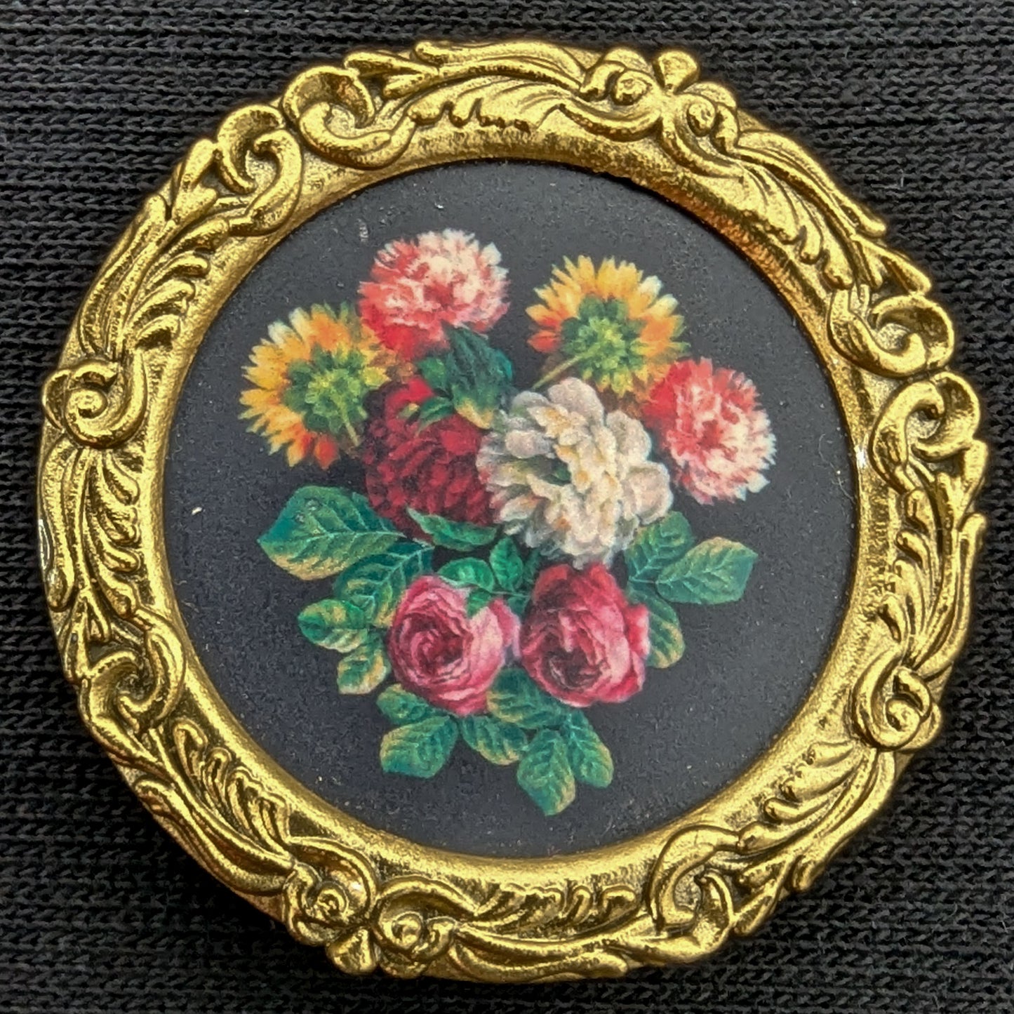 Round gold ornate frame with a miniature floral painting on a black background.