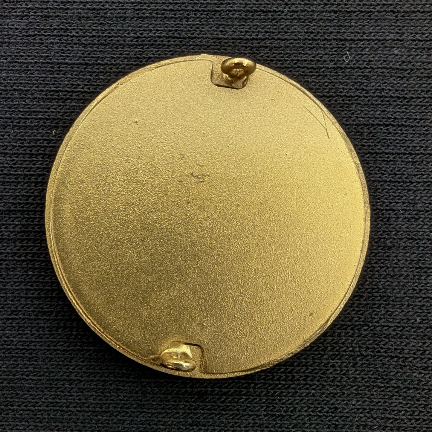 Gold-toned, round D&G Zierelement, 3.5 cm, with two pins on a dark fabric background.