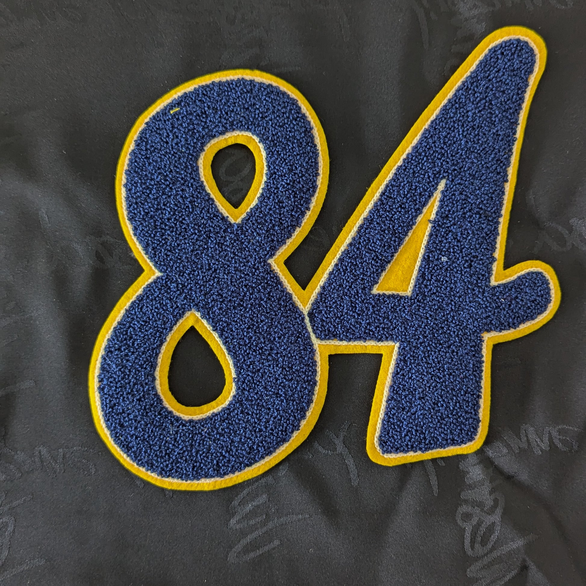 Blue chenille patch with yellow outline, featuring the number "84", on a dark fabric background.