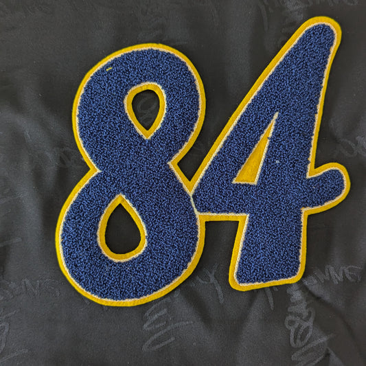 Blue chenille patch with yellow outline, featuring the number "84", on a dark fabric background.