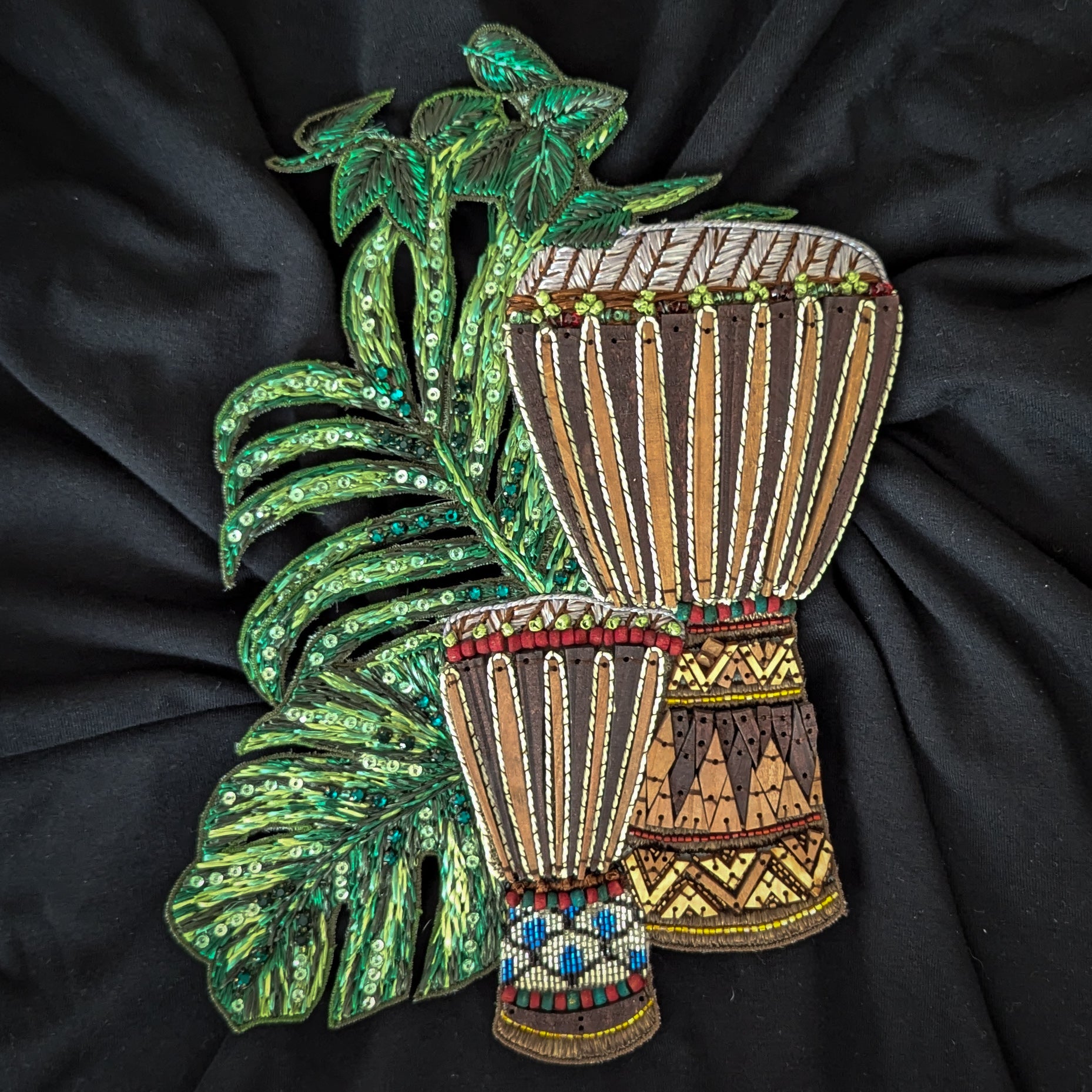 Embroidered patch with two brown drums, green sequined leaves, and colorful bead details on black fabric.