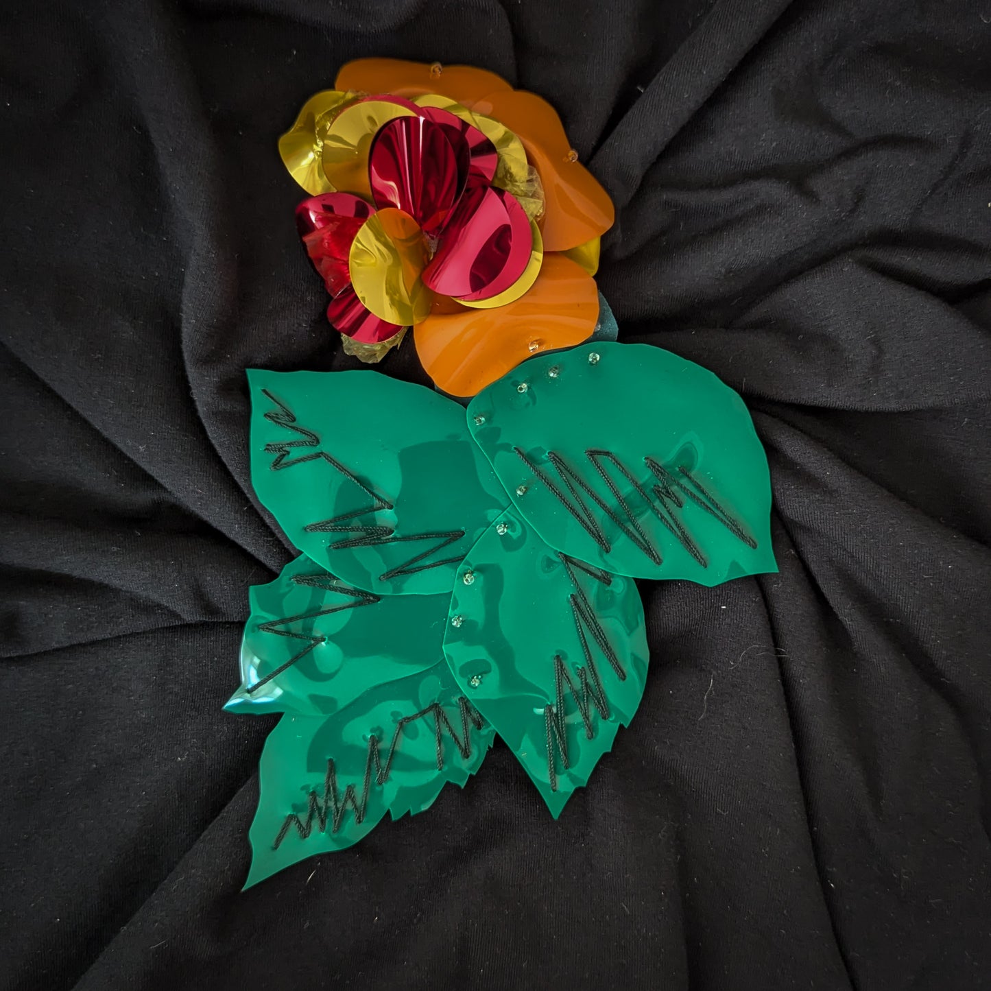 D&G plastic flower patch with shiny red, orange, yellow petals and green leaves on black fabric.
