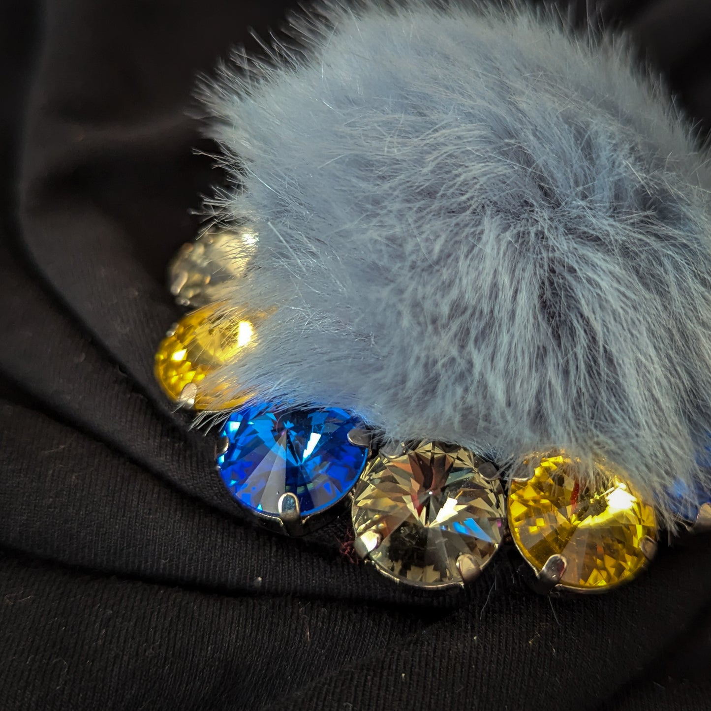 Light blue faux fur ornament with colorful Swarovski crystals on black fabric.