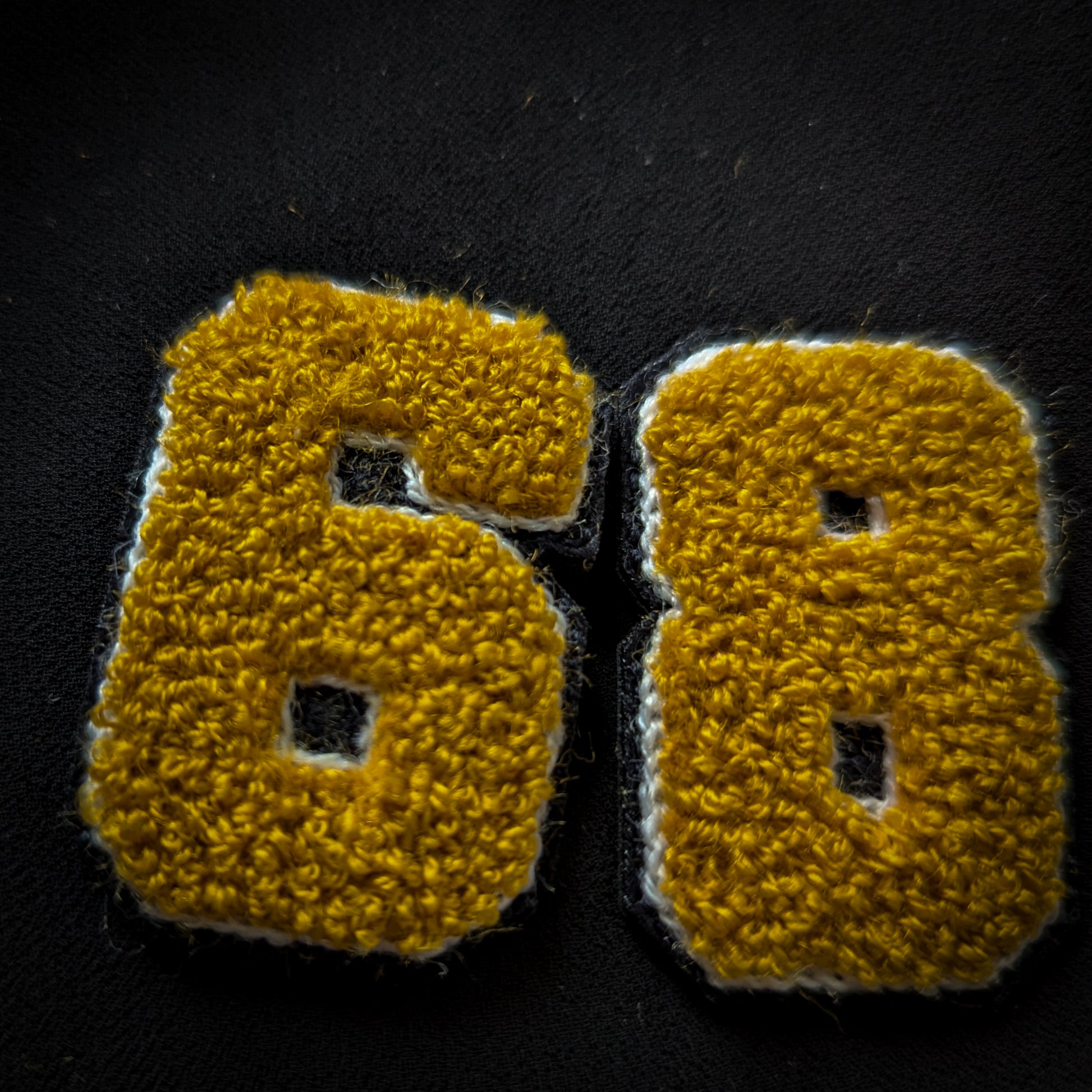 D&G gold chenille patch with "68" numbers, outlined in white and black, on a dark fabric.