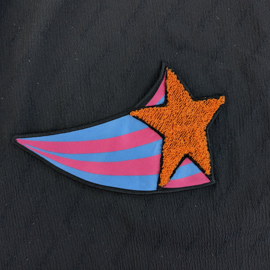 Orange embroidered star patch with pink and blue striped tail on black fabric.