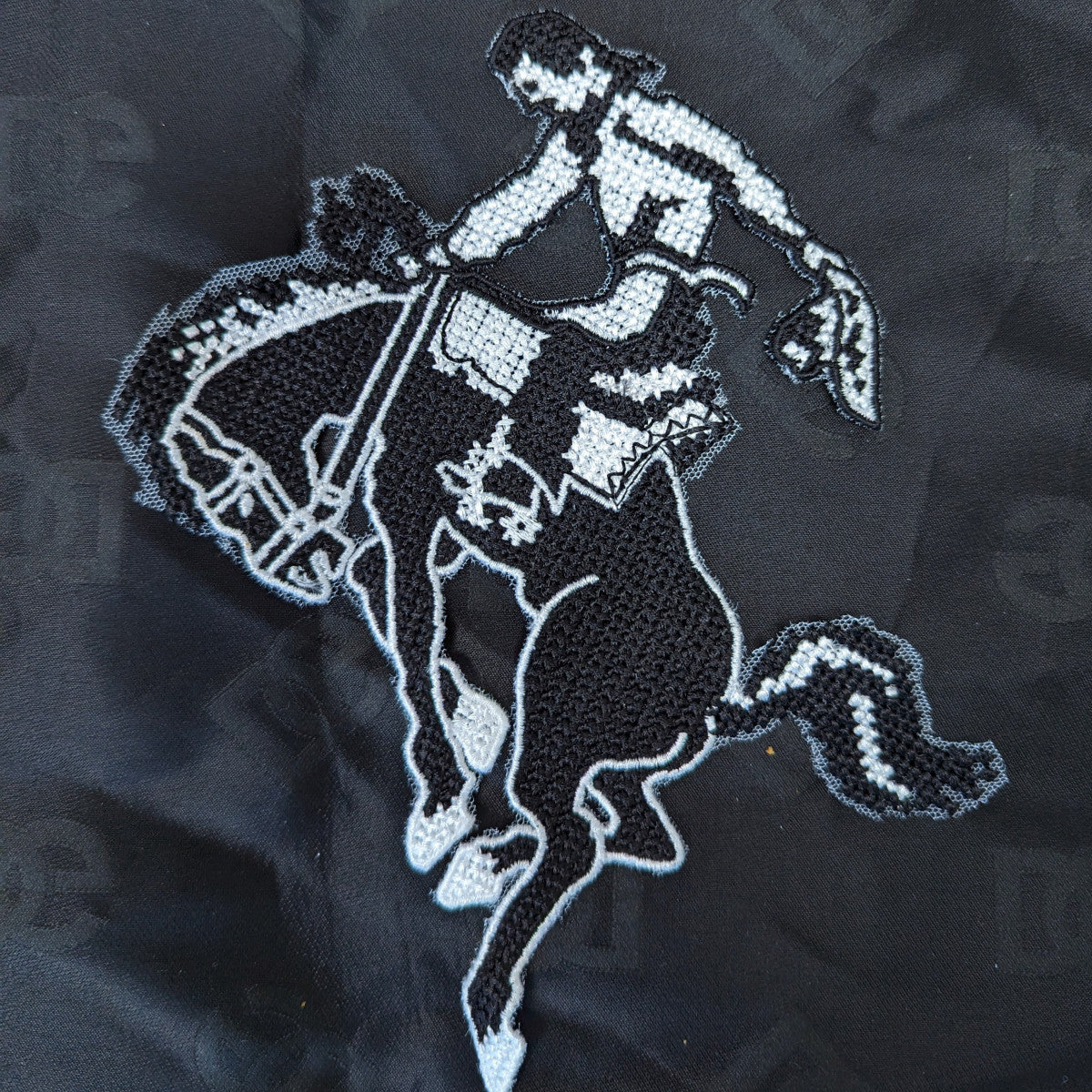 Black and white embroidered cowboy riding a bucking horse patch, 15x20cm, on a black fabric background.