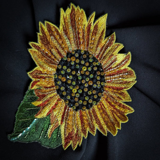 Embroidered sunflower patch with yellow, orange, and brown petals, beaded center, and green leaf.