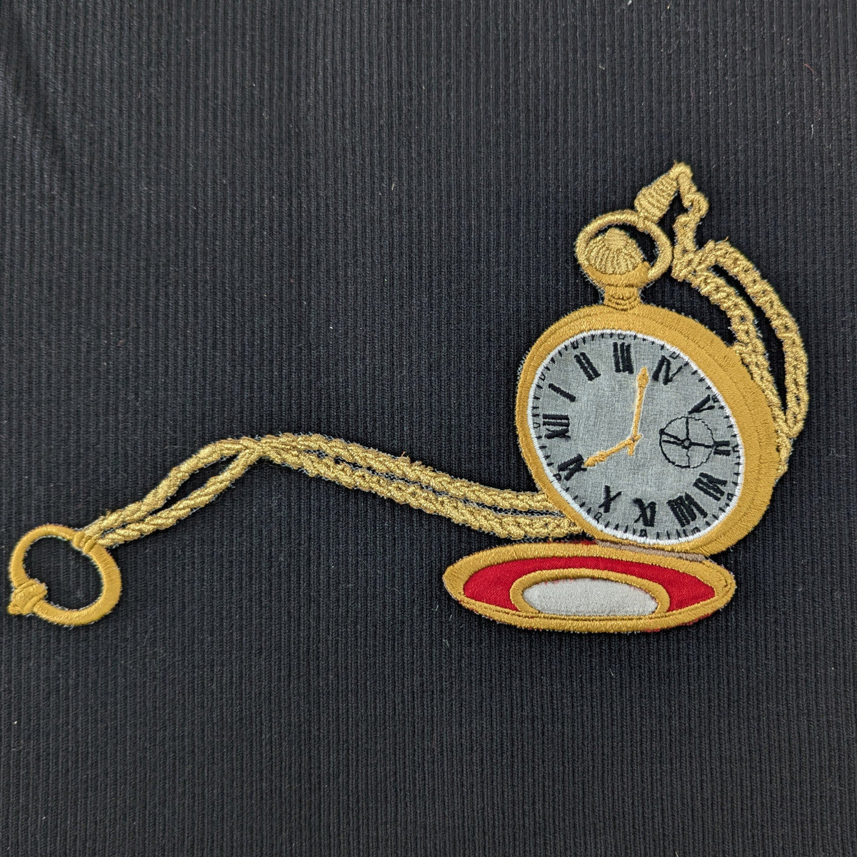 Embroidered gold pocket watch patch with chain and red stand on black fabric.
