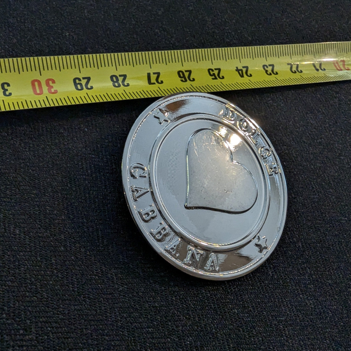 Shiny silver Dolce & Gabbana button, 4.5 cm, with a central heart design and stars, next to a measuring tape.
