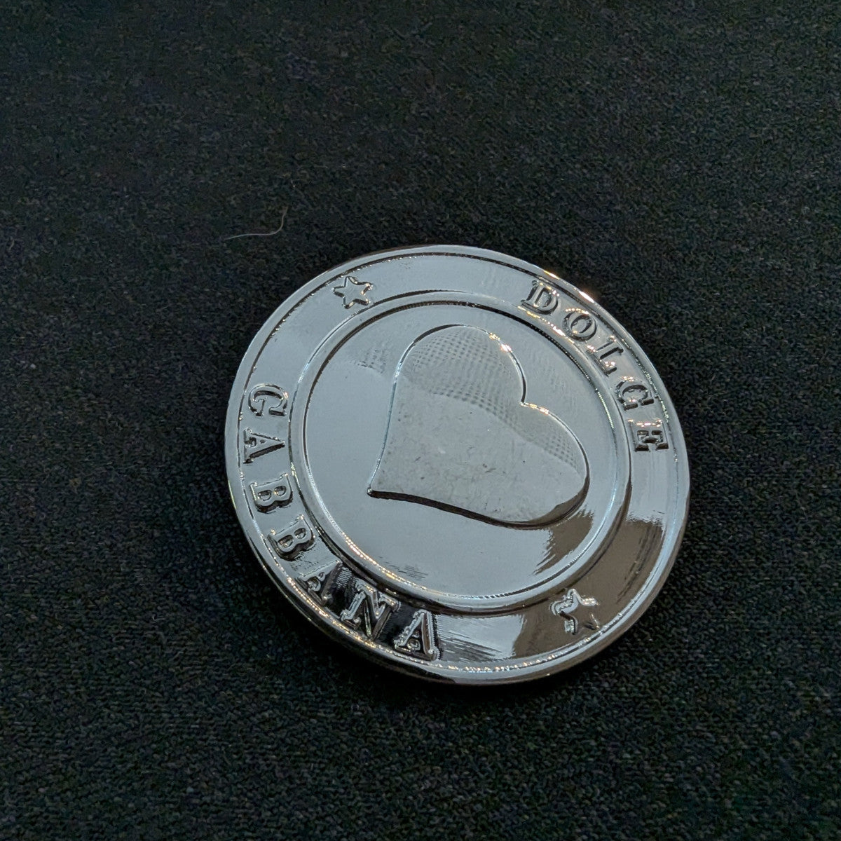 Shiny silver Dolce & Gabbana designer button, 4.5 cm, with a raised heart and stars on black fabric.