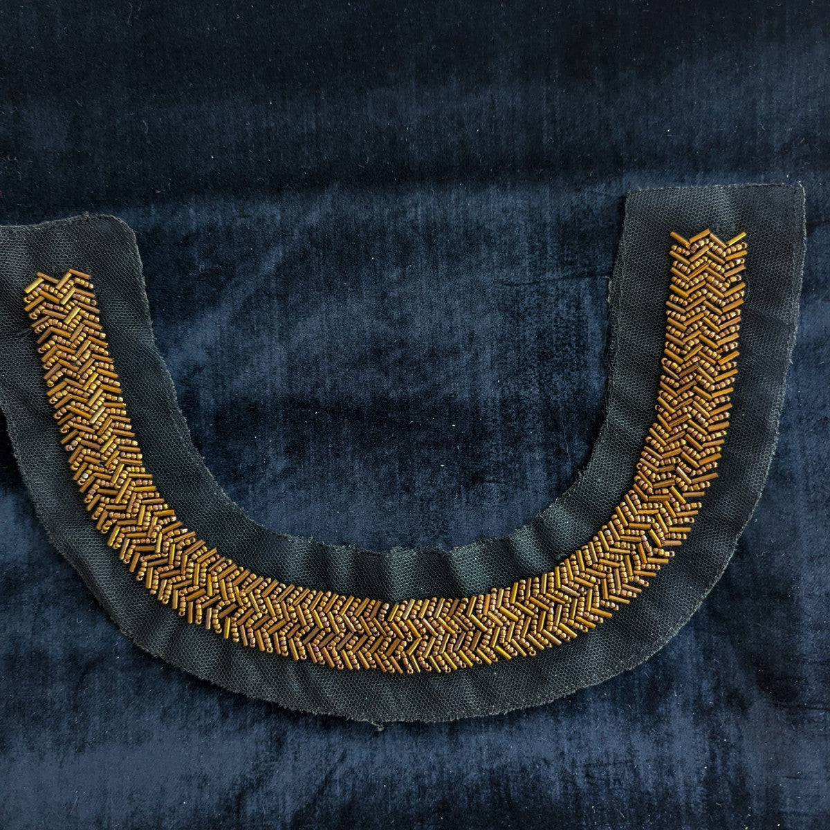 Black fabric patch with U-shaped gold and bronze beaded embroidery in a chevron pattern.
