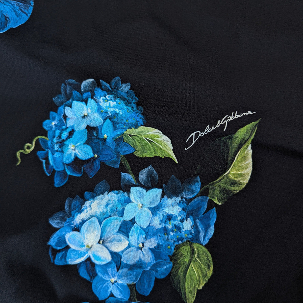 Black silk fabric with vibrant blue hydrangea floral print and "Dolce&Gabbana" logo.