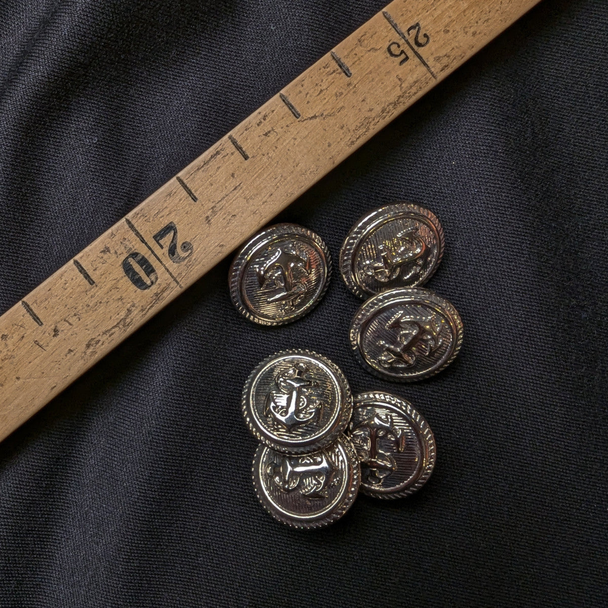 Six 2cm silver metal buttons with anchor design on black fabric next to a wooden ruler.