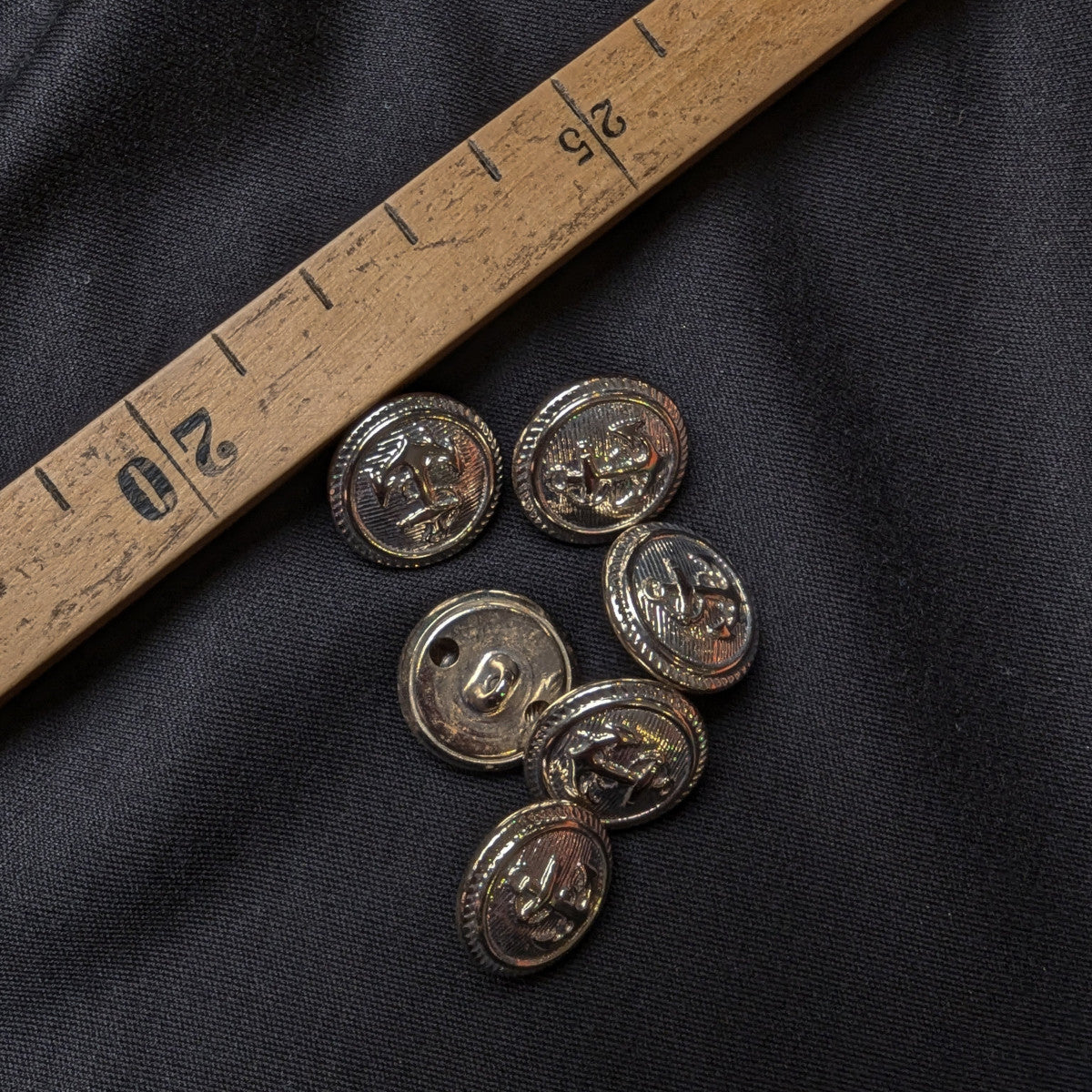 Six 2cm silver metal buttons with an embossed design on black fabric, next to a wooden ruler.