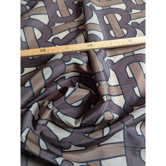 Brown and beige B*rberry deadstock polyester fabric with all-over logo, shown with a ruler.