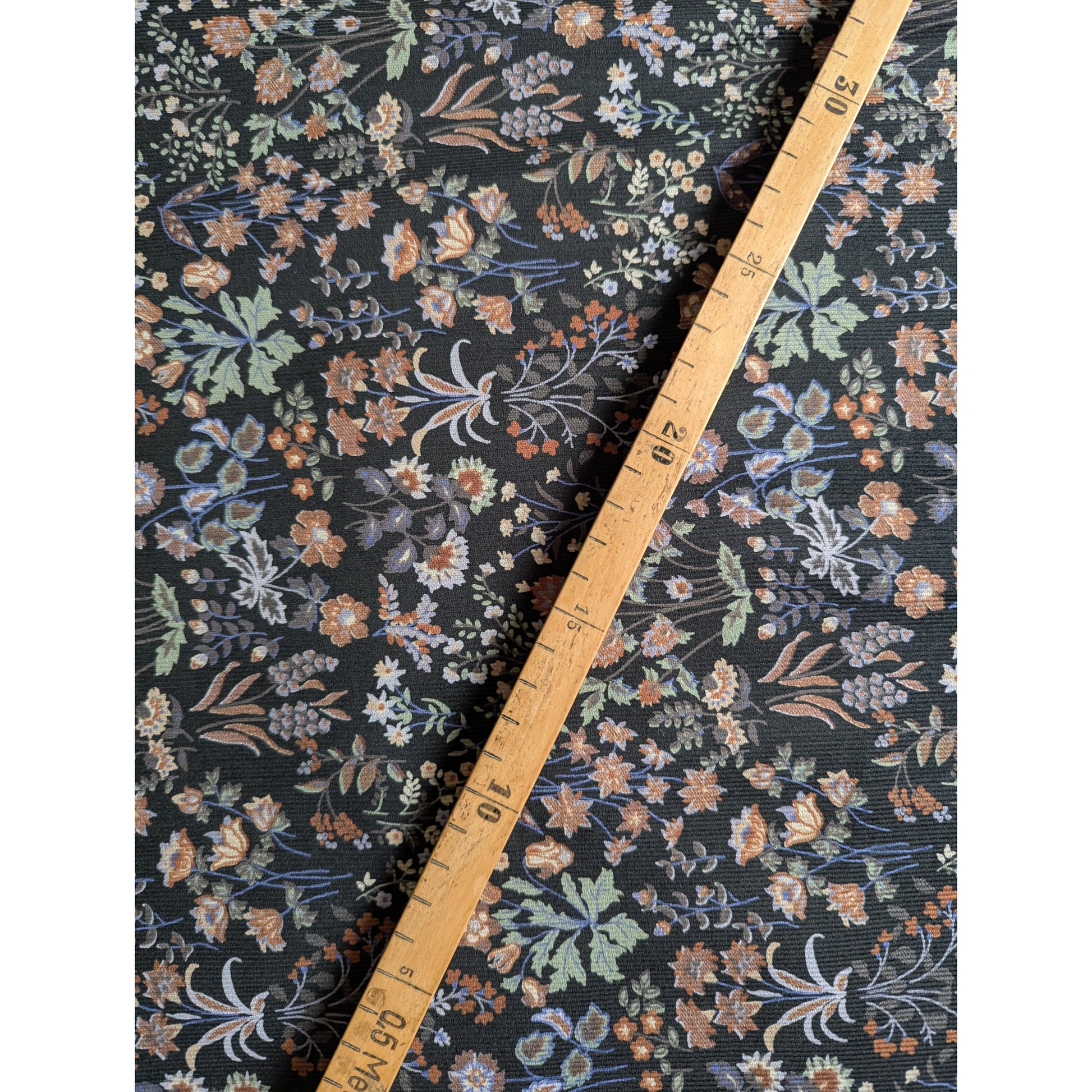 Black Feincord fabric with a vibrant multicolor millefleur floral pattern, measured by a wooden ruler.