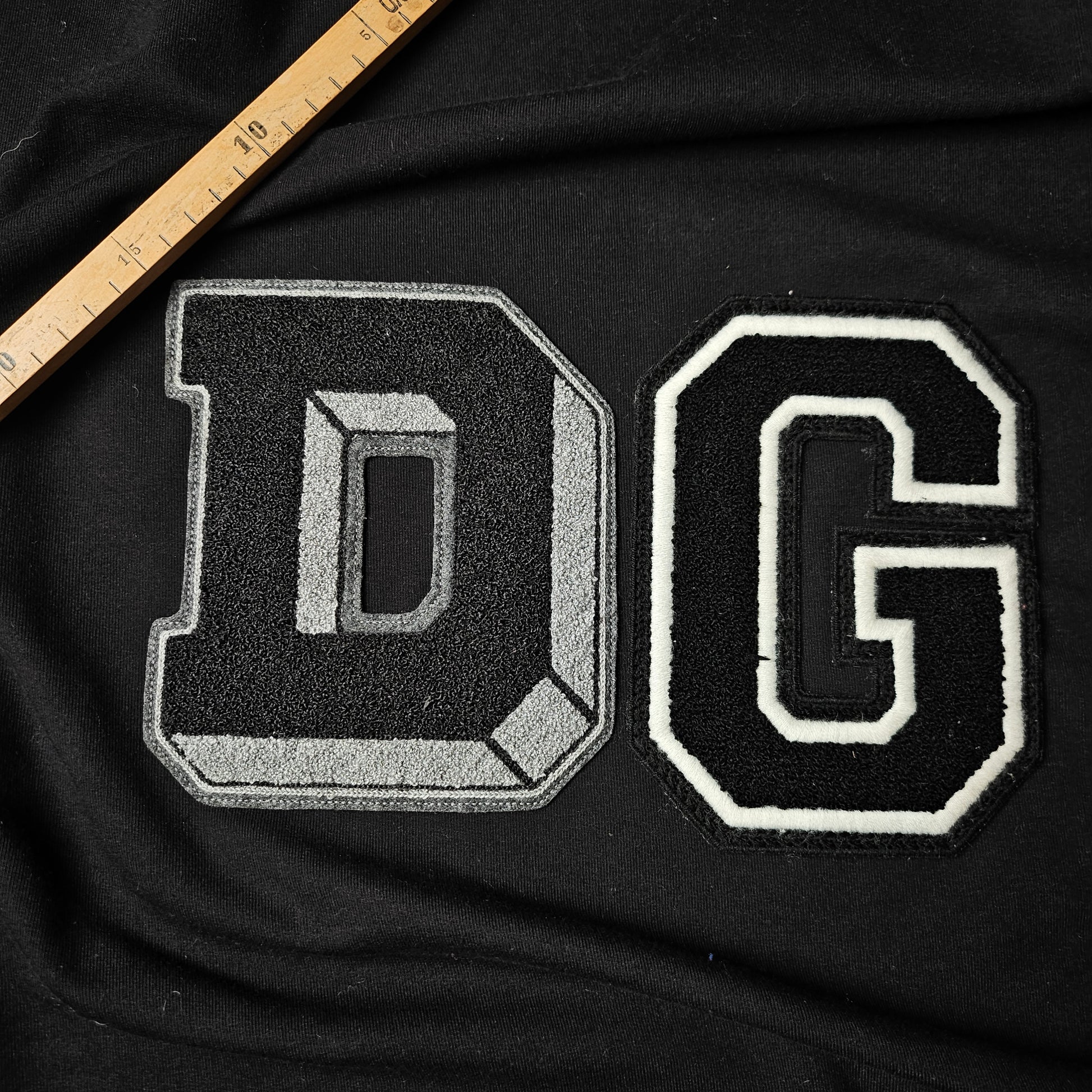 Black and gray DG patch, 15x15cm, with white outline on black fabric, next to a wooden ruler.