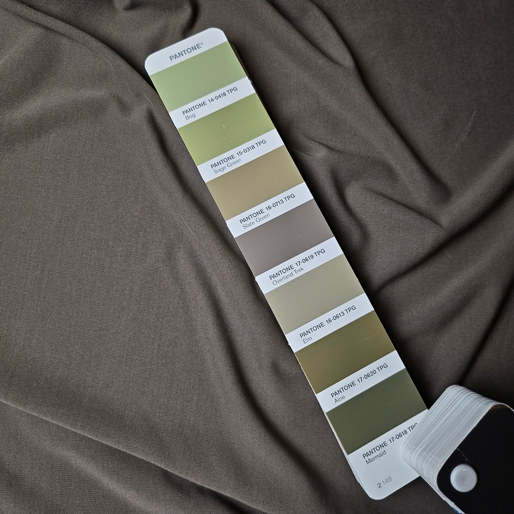 Finely textured mud Viskosejersey Crepe fabric, draped, with a Pantone fan highlighting "Overland Trek" greens.