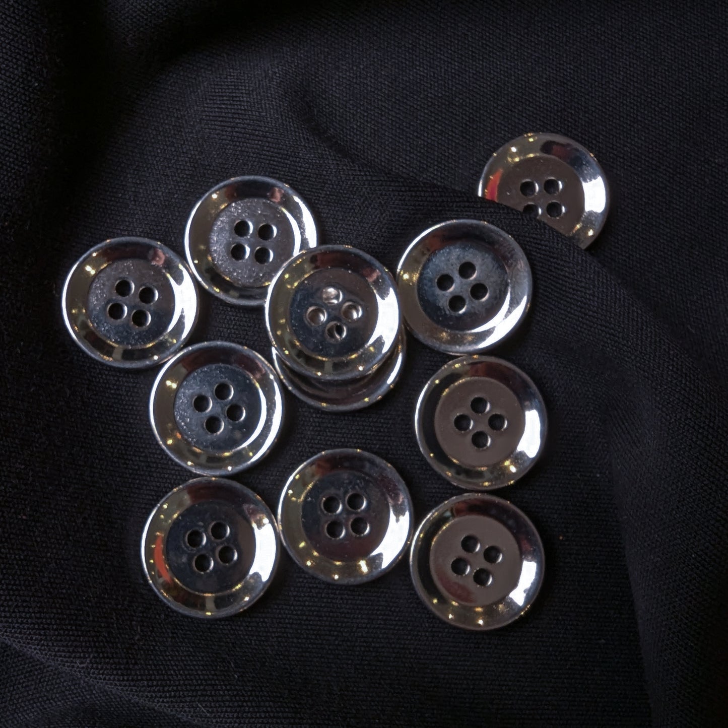 Ten shiny silver 1.5 cm metal 4-hole buttons on black fabric.