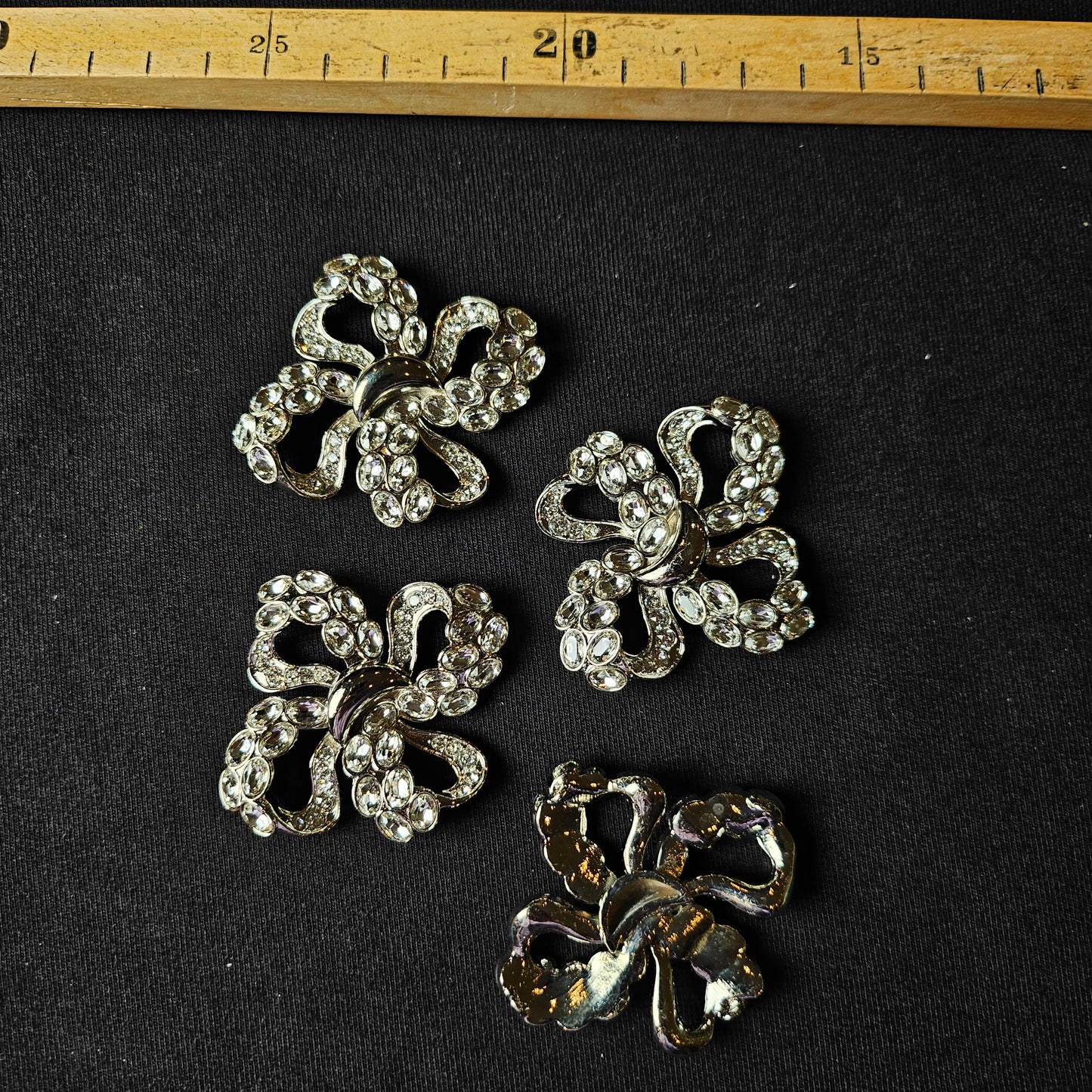 Four silver D&G flower ornaments with sparkling Swarovski stones on black fabric, with a ruler above.