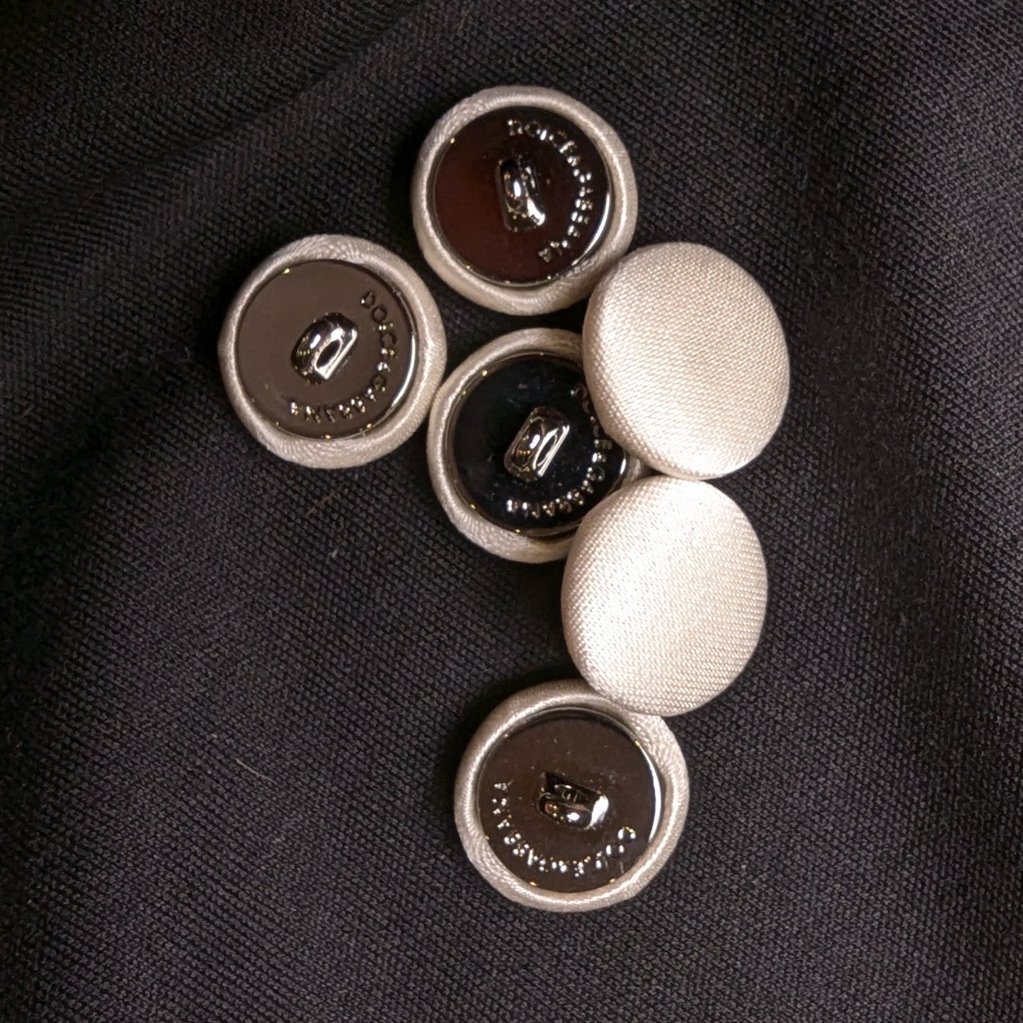 Set of six 1.5cm ivory silk-covered D&G buttons with metal backs, on black fabric.