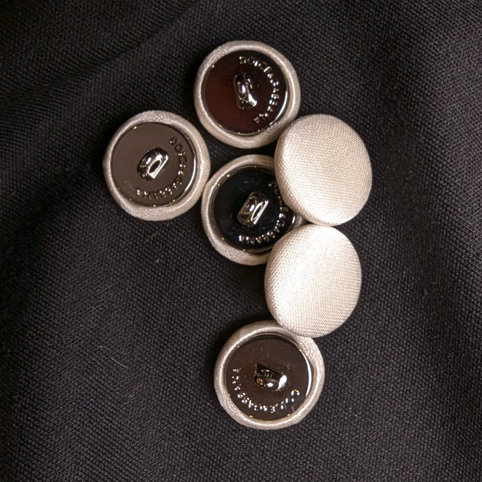 Set of six 1.5cm ivory silk-covered D&G buttons with metal backs, on black fabric.